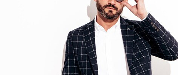 Стоит мужчина татуированный с бородой в клетчатом пиджаке с белой рубашкой презентует формальный стиль